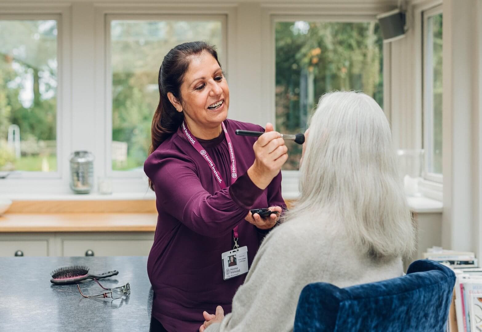 A caregiver in a purple shirt applies makeup to an elderly woman with white hair in a cozy, sunlit room. - Home Instead