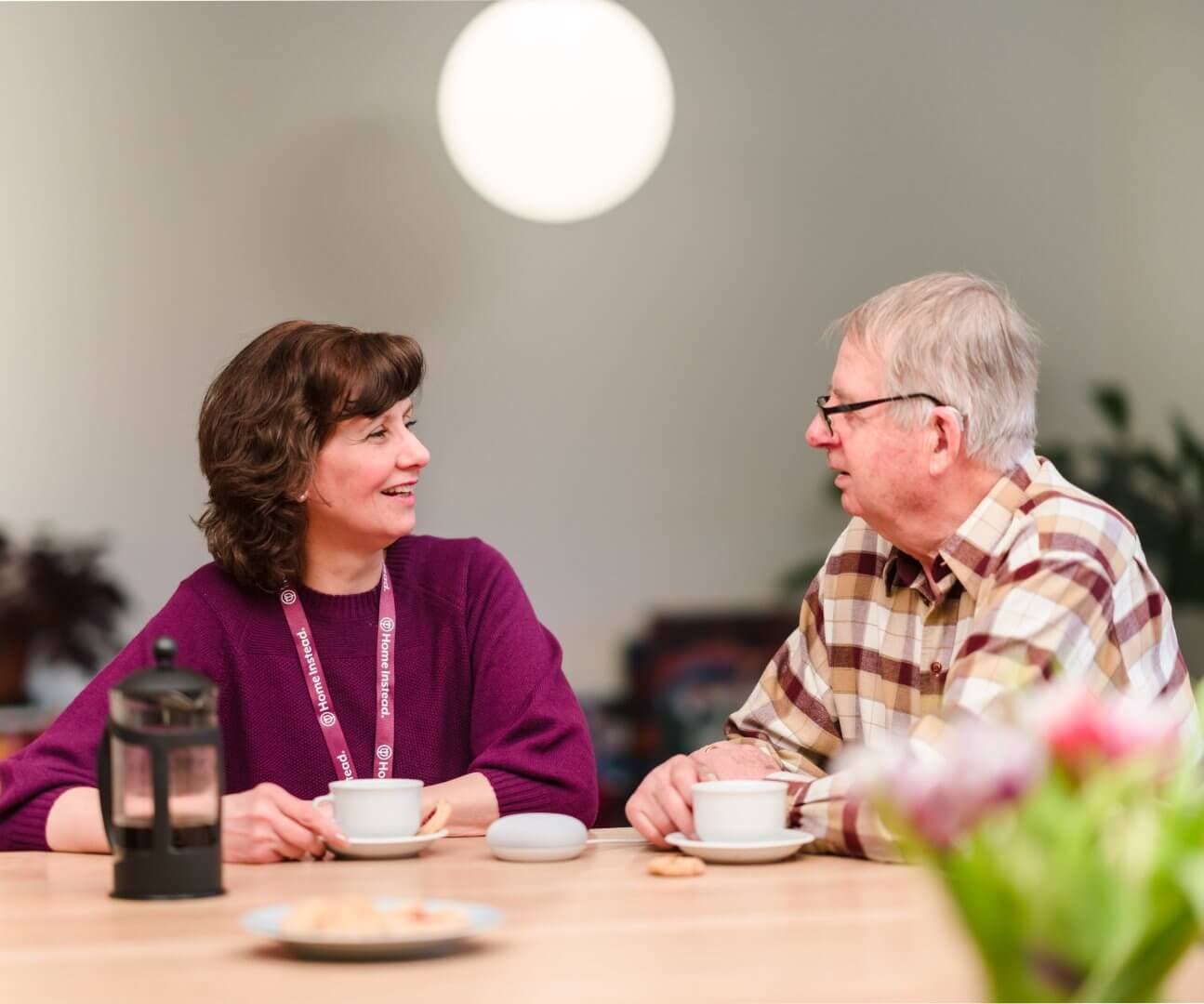 A woman and an older man sit at a table with coffee cups, smiling and engaging in conversation. - Home Instead