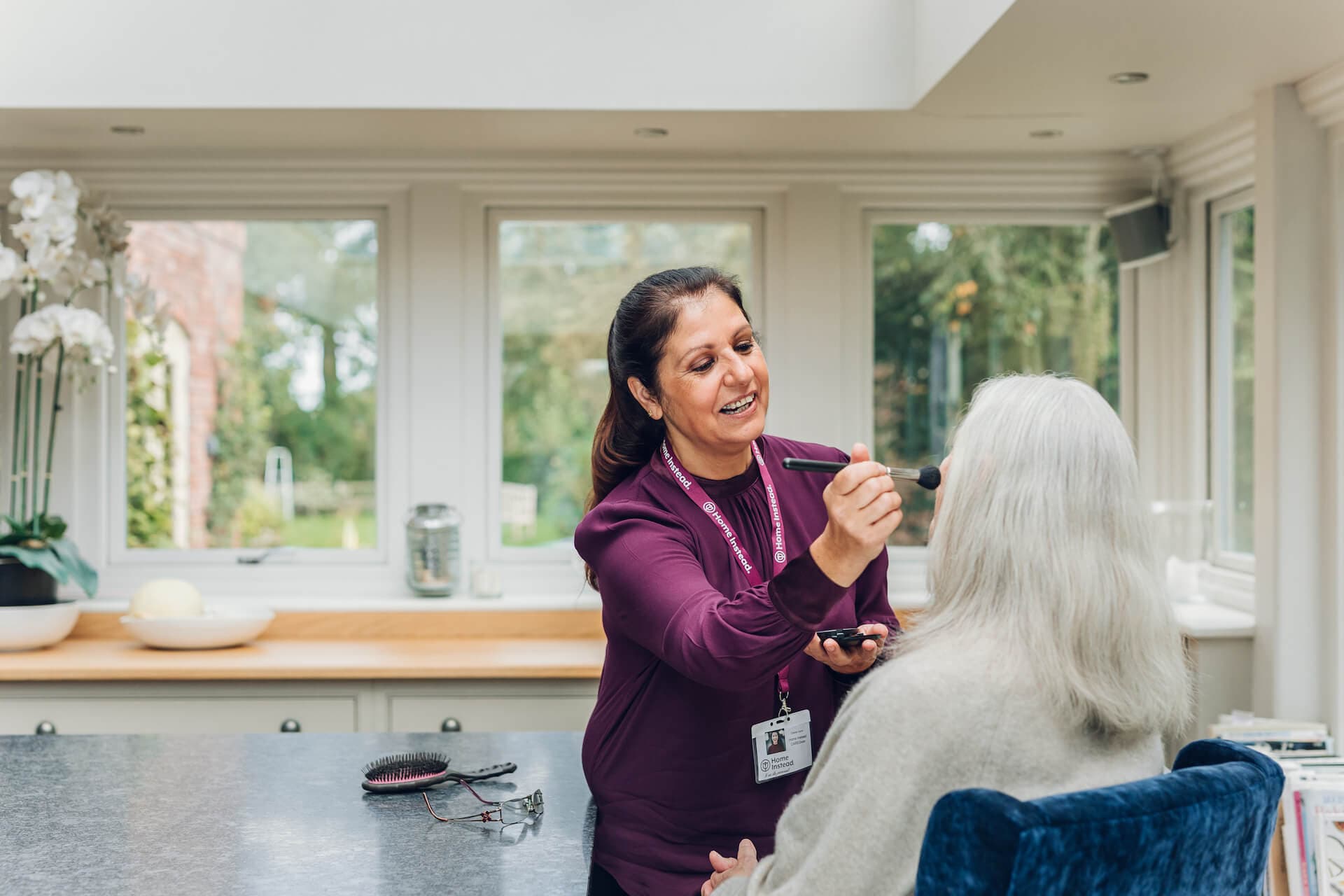 A woman applies makeup to an elderly woman's face in a bright, modern room with large windows. - Home Instead