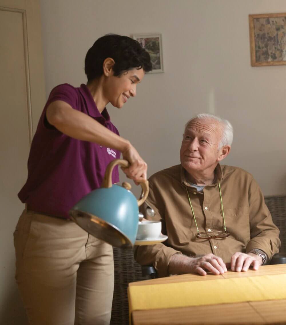 A caregiver wearing a mask tends to an elderly man sitting in a wheelchair next to a window with sheer curtains. - Home Instead