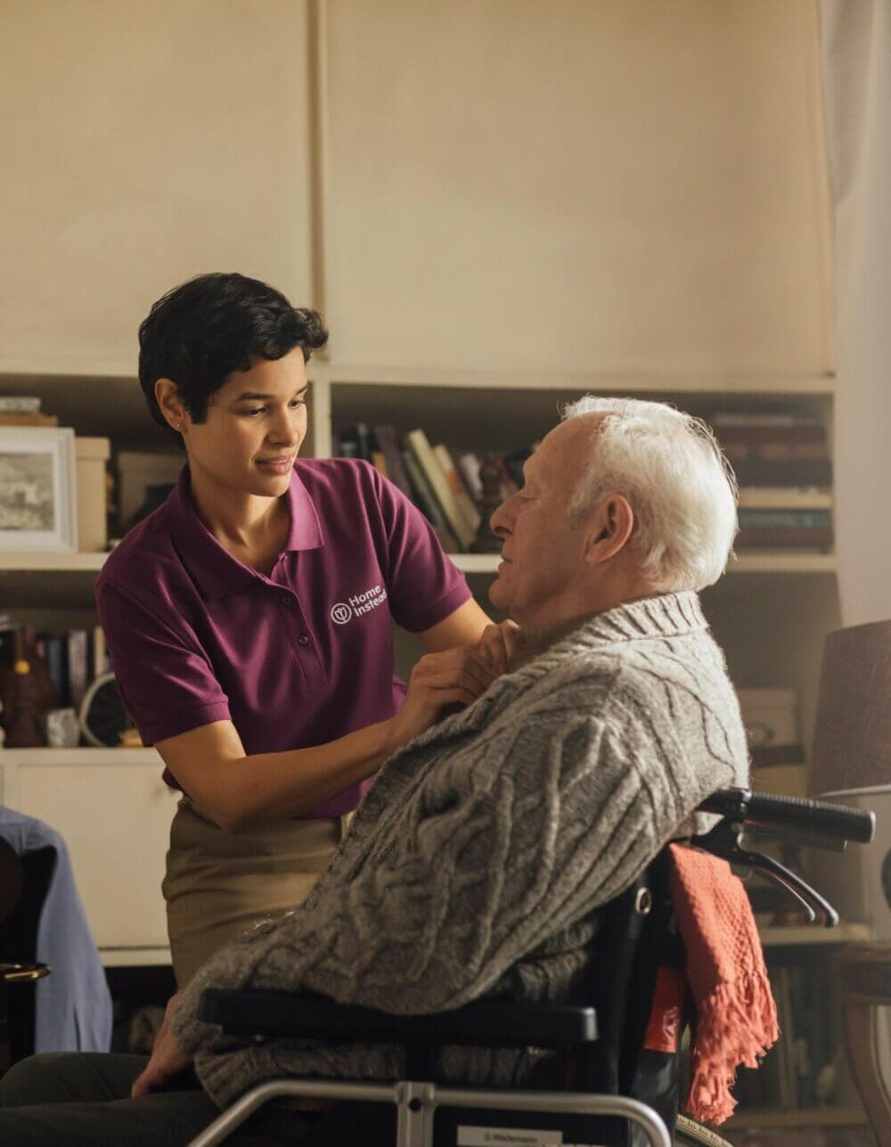 An elderly man in a wheelchair smiles as a healthcare worker in a mask checks his ears in a softly lit room. - Home Instead