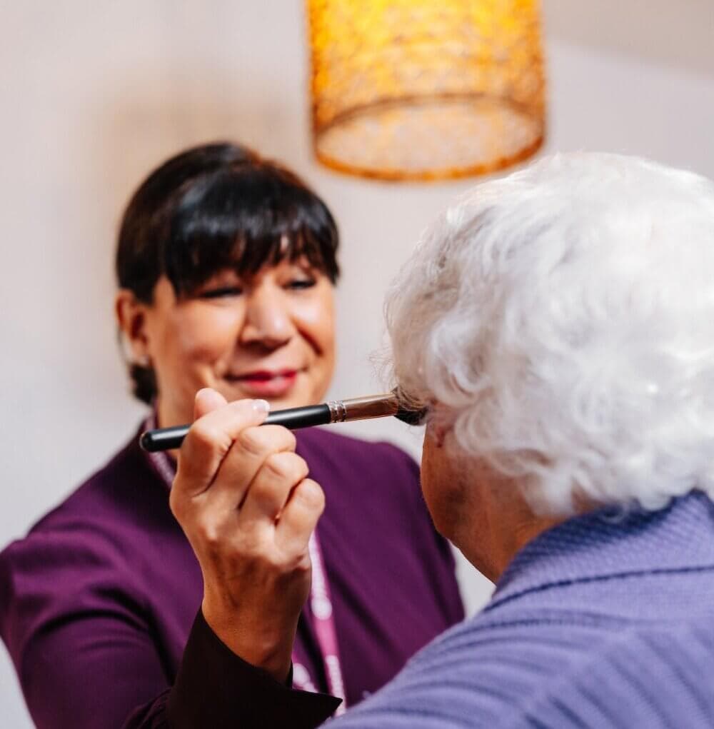 A caregiver in a purple polo shirt assists an elderly man in a wheelchair with adjusting his sweater. - Home Instead