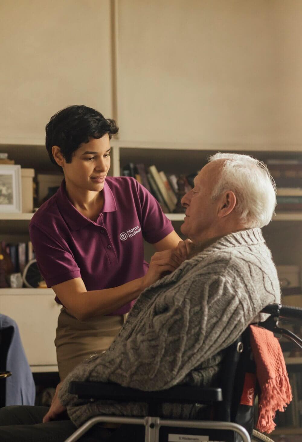 A caregiver assists an elderly person in a wheelchair, adjusting their sweater in a cozy, homey room. - Home Instead