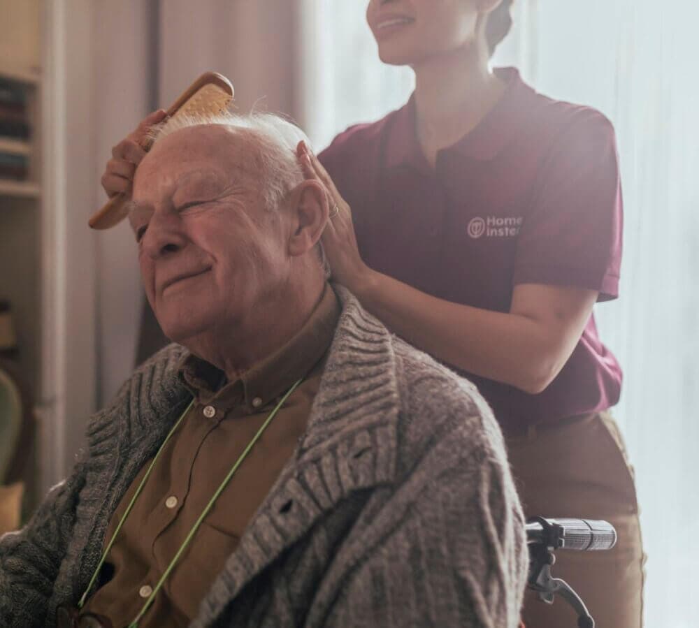 Caregiver in a purple shirt assists a seated elderly person in a wheelchair in a cozy room. - Home Instead