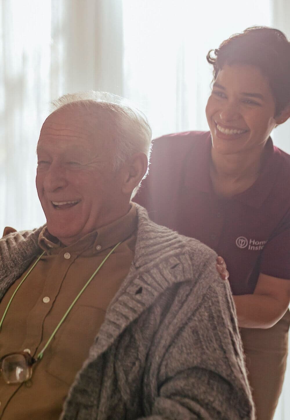 An older man in a knit sweater laughs with a caregiver in a maroon shirt standing behind him, both looking happy. - Home Instead