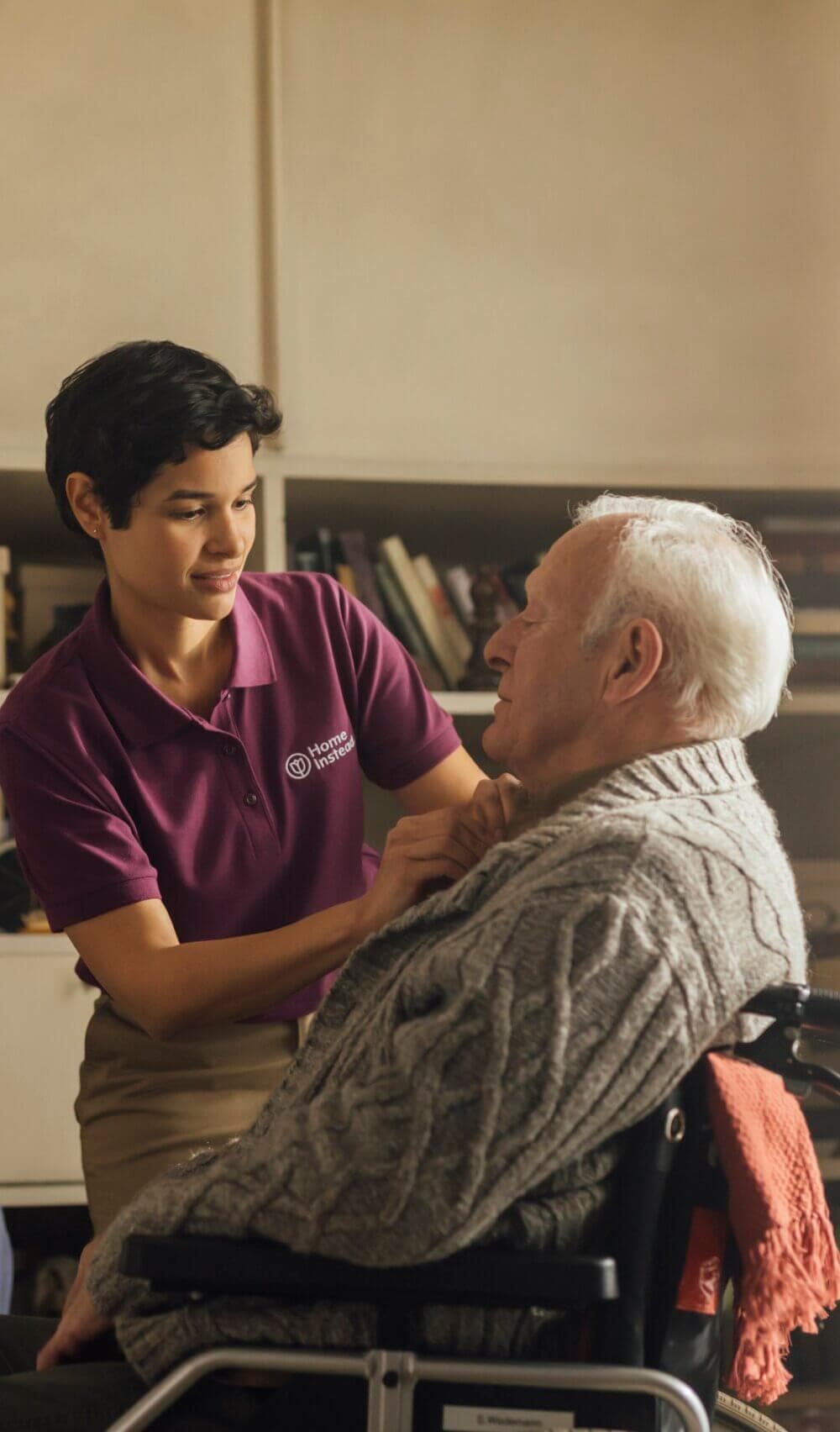 A caregiver in a purple shirt helps an elderly man in a wheelchair adjust his sweater in a cozy room. - Home Instead