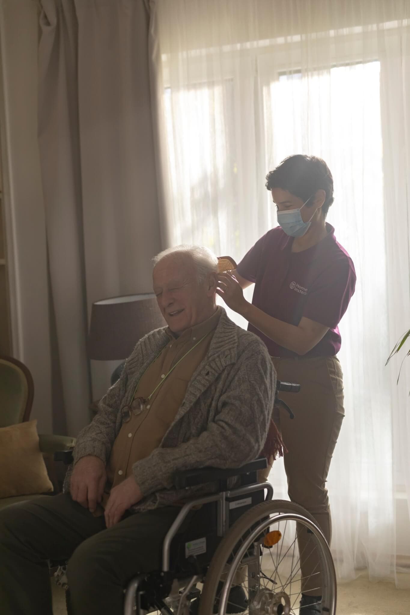 A caregiver wearing a mask assists an elderly man in a wheelchair with grooming, in a softly lit room. - Home Instead