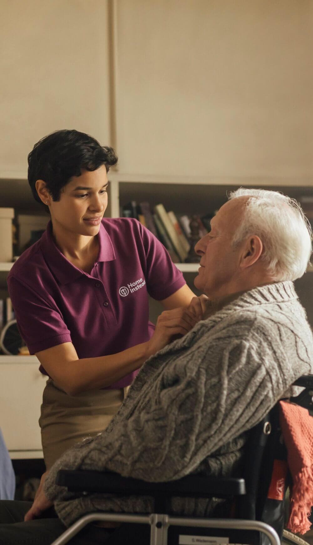 A caregiver wearing a purple shirt assists an elderly man in a wheelchair with adjusting his sweater. - Home Instead