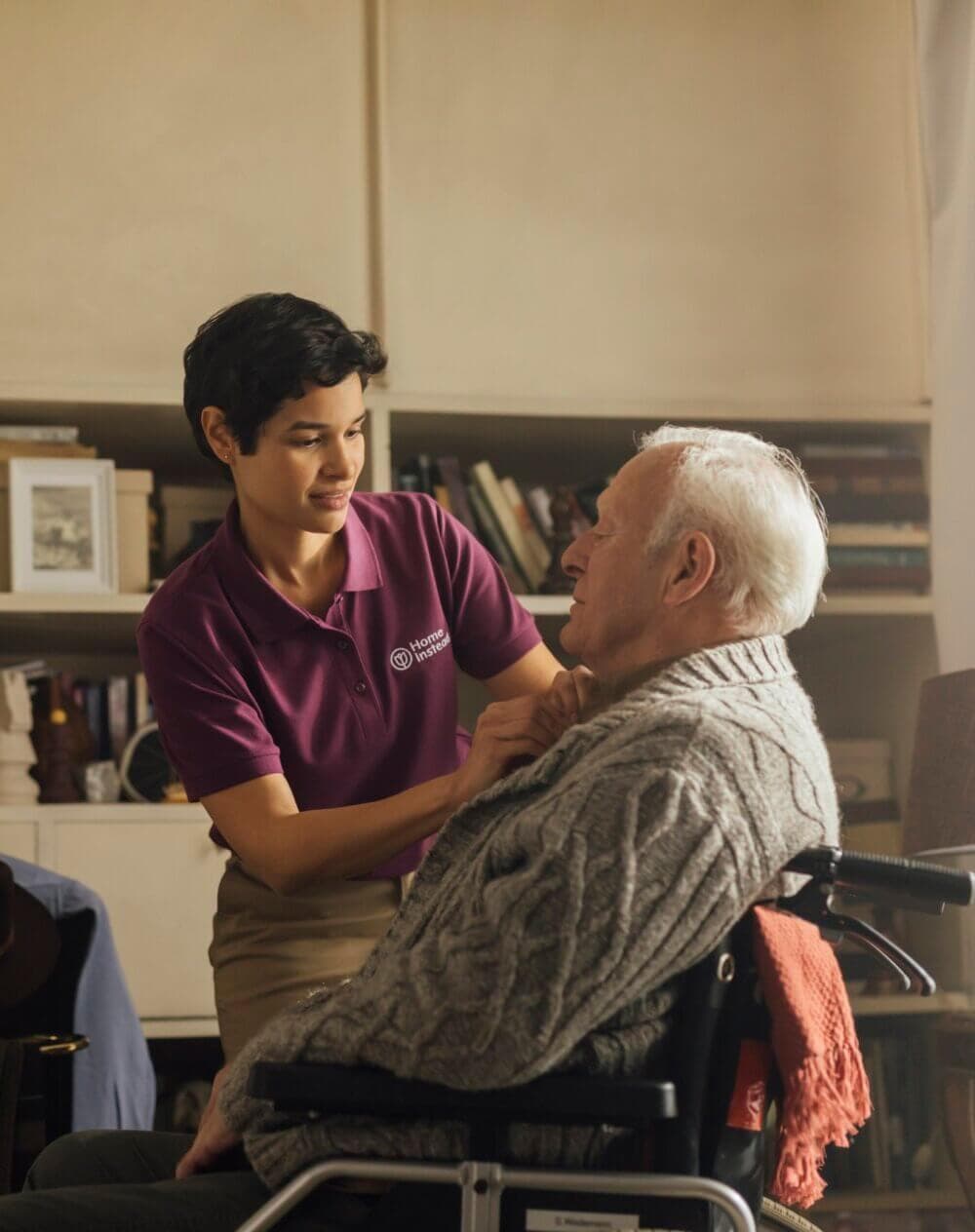 A caregiver helps an elderly person in a wheelchair by adjusting their sweater in a cozy, homey room. - Home Instead