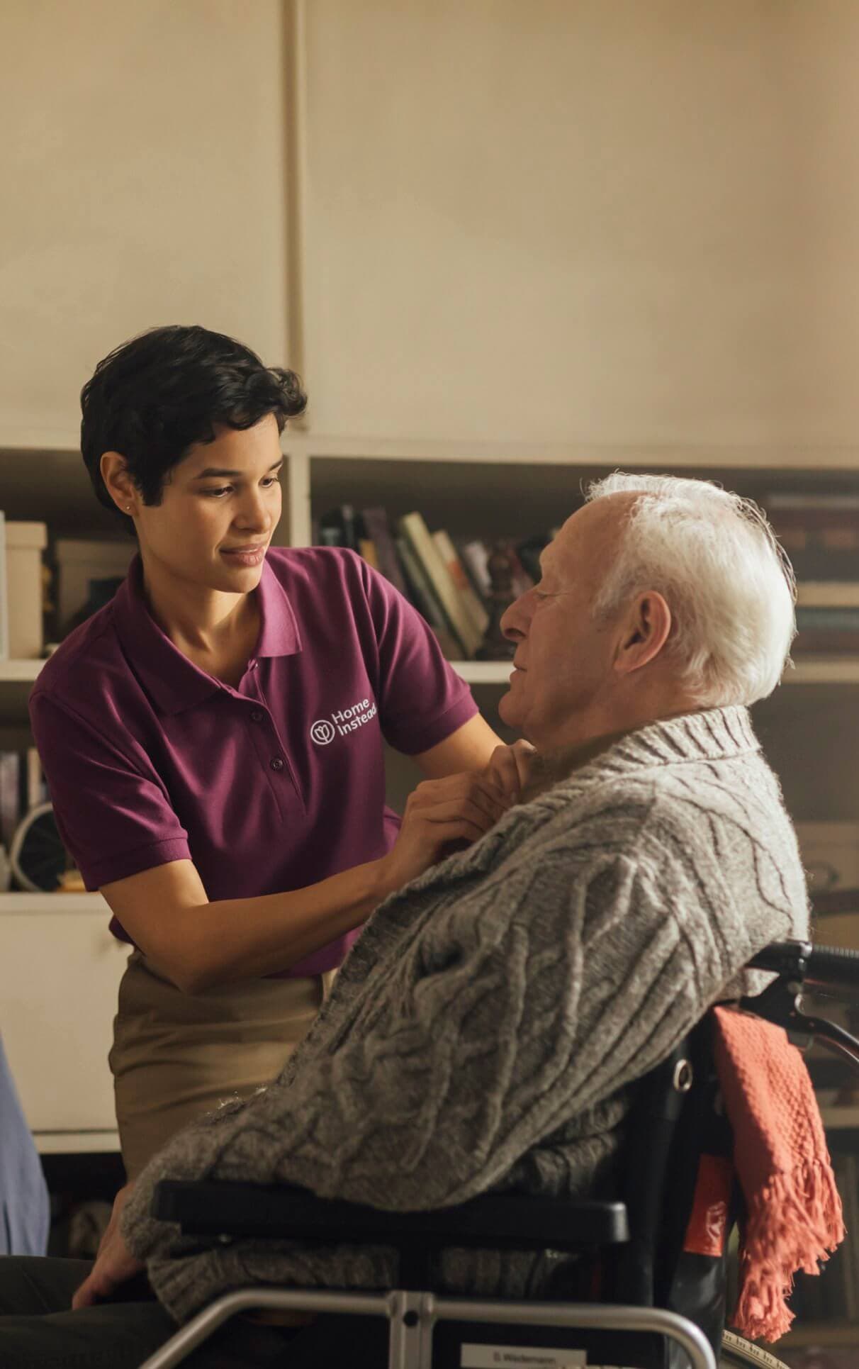 A caregiver in a purple shirt assists an elderly man in a wheelchair by buttoning his sweater. - Home Instead