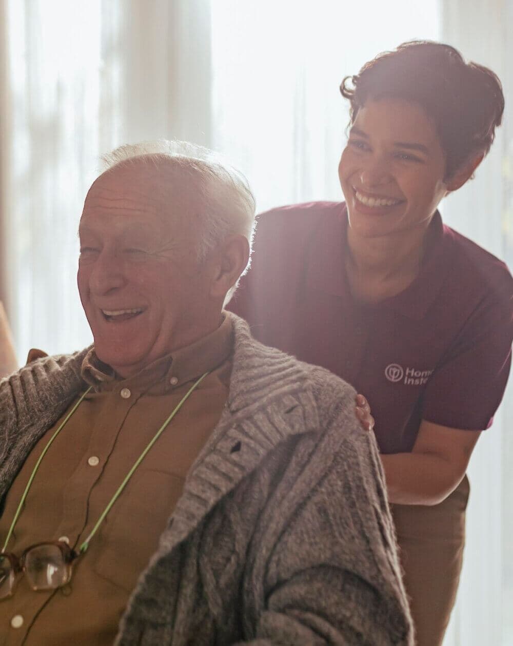 An elderly man with glasses laughs with a Home Instead Poole Care Professional, smiling behind him, in a warmly lit room.