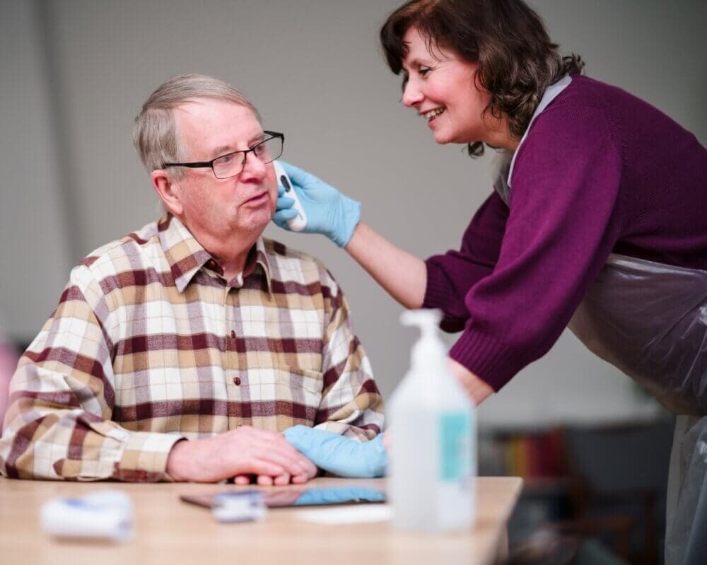 A caregiver in a purple uniform assists an elderly man in a wheelchair indoors. - Home Instead