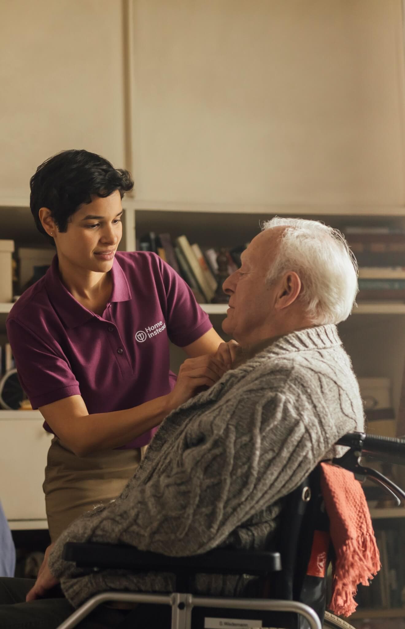 A caregiver in a purple uniform assists an elderly man in a wheelchair indoors. - Home Instead