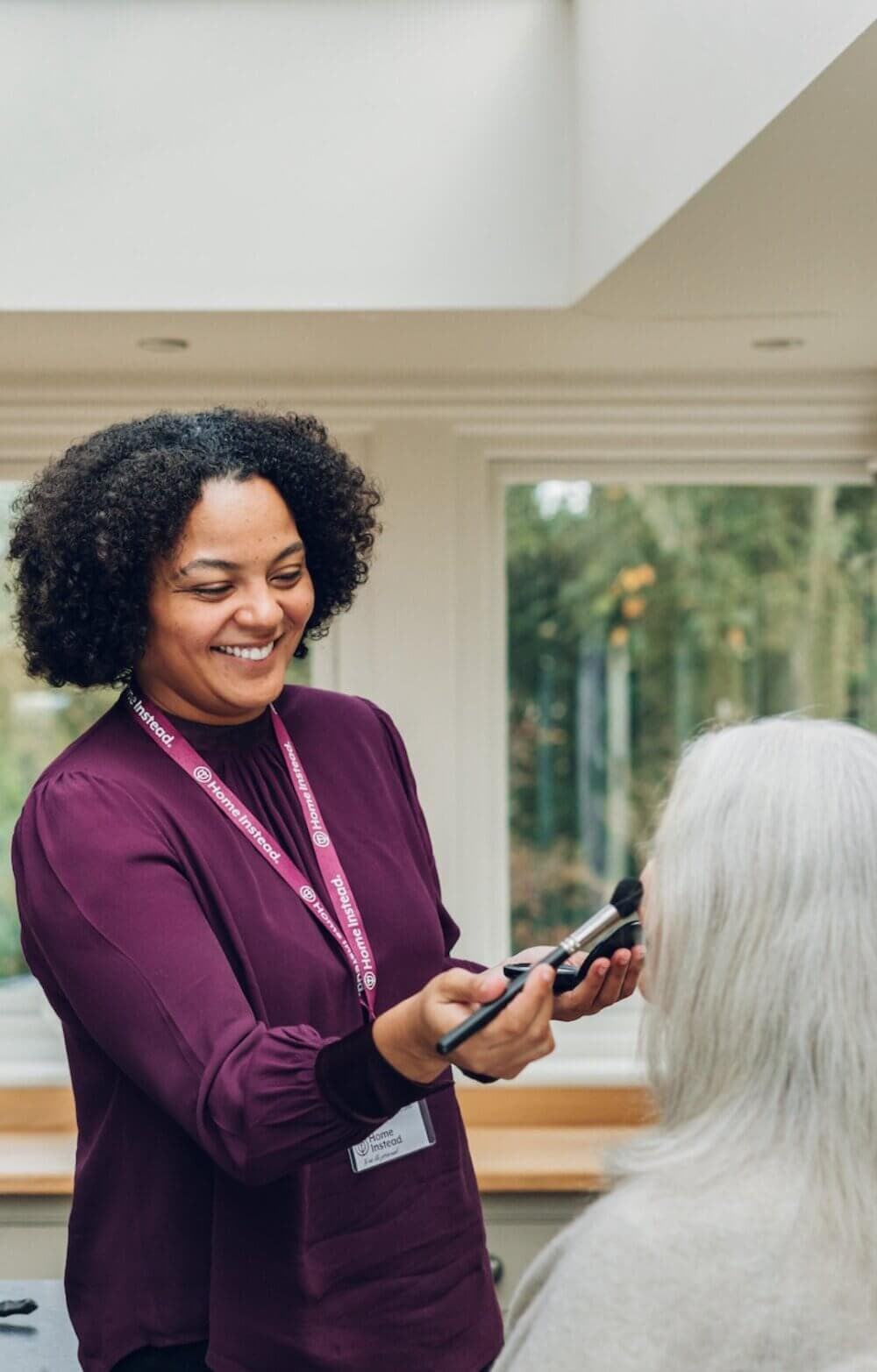 A woman with curly hair smiles as she applies makeup to the face of an older person with long white hair. - Home Instead