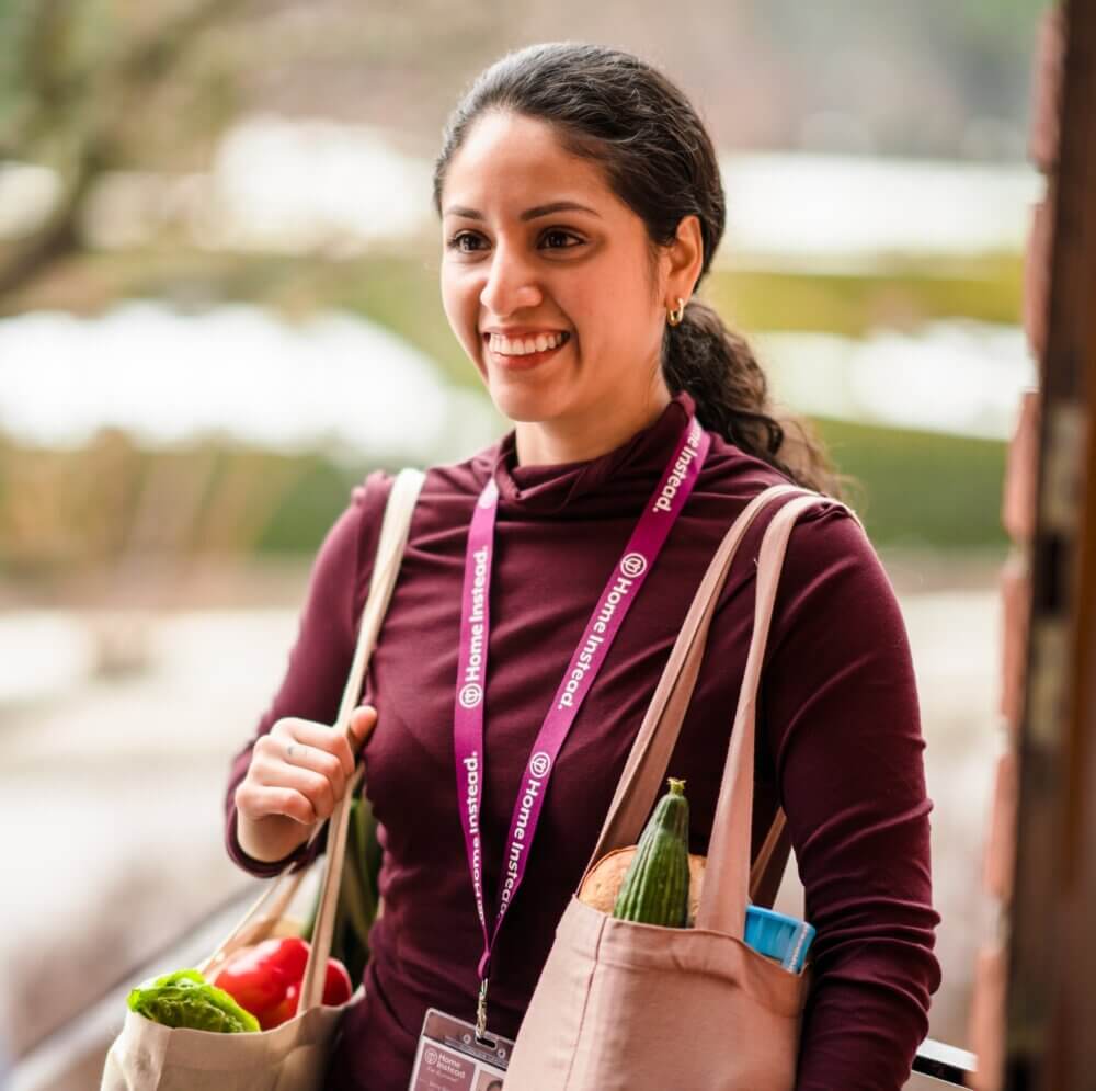 A smiling woman in a maroon blouse applies makeup to an older woman with long white hair. - Home Instead