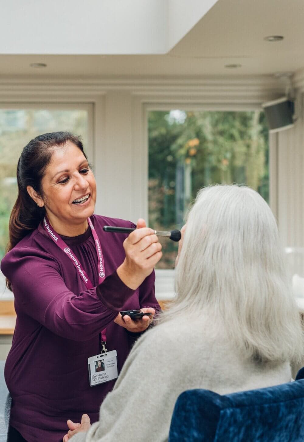 A caregiver applying makeup to a senior woman seated in a well-lit room with large windows. - Home Instead