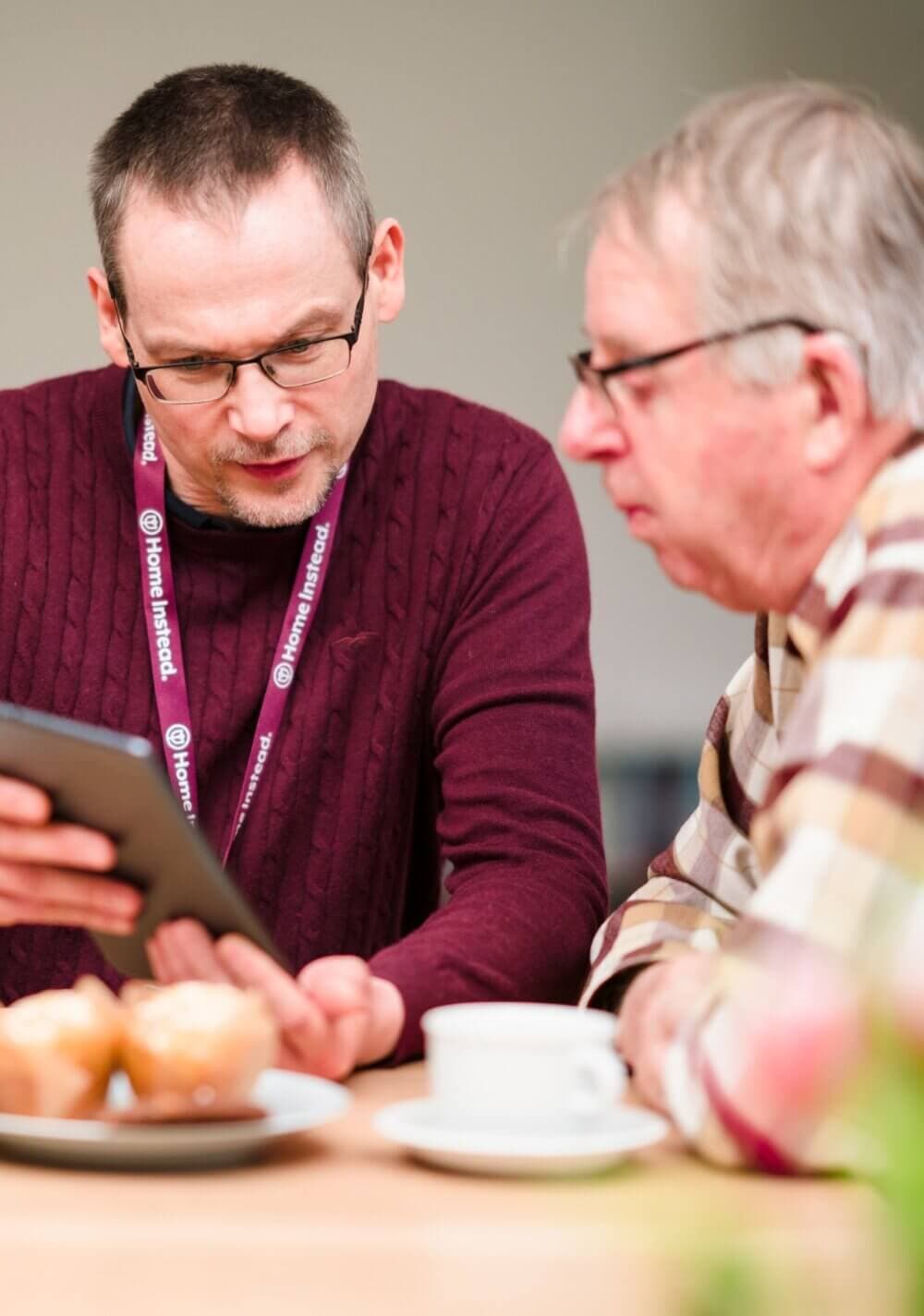 Two men looking at a tablet, one wearing glasses and a maroon sweater, the other in a plaid shirt, sitting at a table. - Home Instead