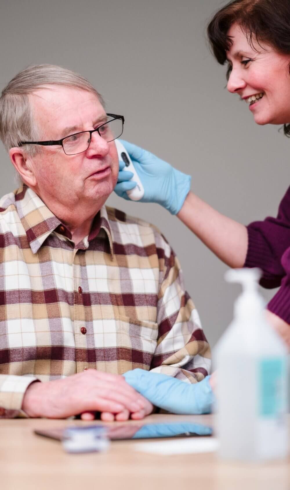 A woman wearing gloves gently wipes an older man’s face as he sits, both smiling. - Home Instead