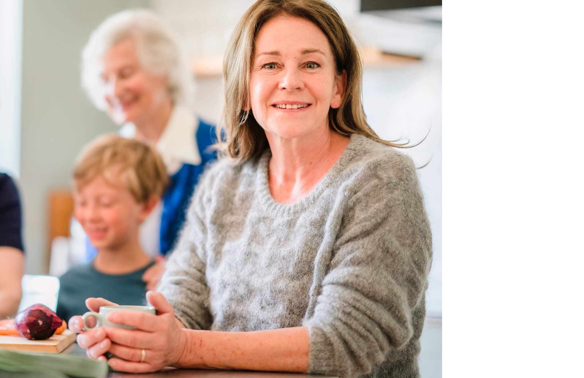 A woman with long hair smiles while seated with a child and an elderly woman in the background. - Home Instead