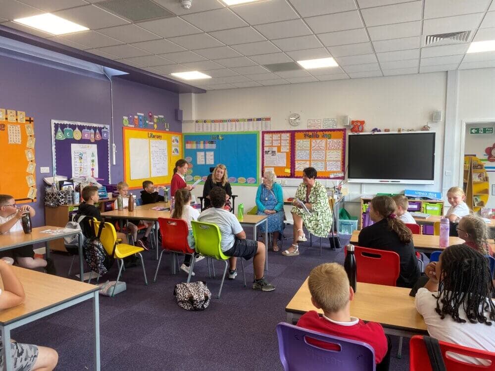 Classroom with students sitting at desks, four adults at the front engaged in discussion, and colorful posters on purple walls. - Home Instead