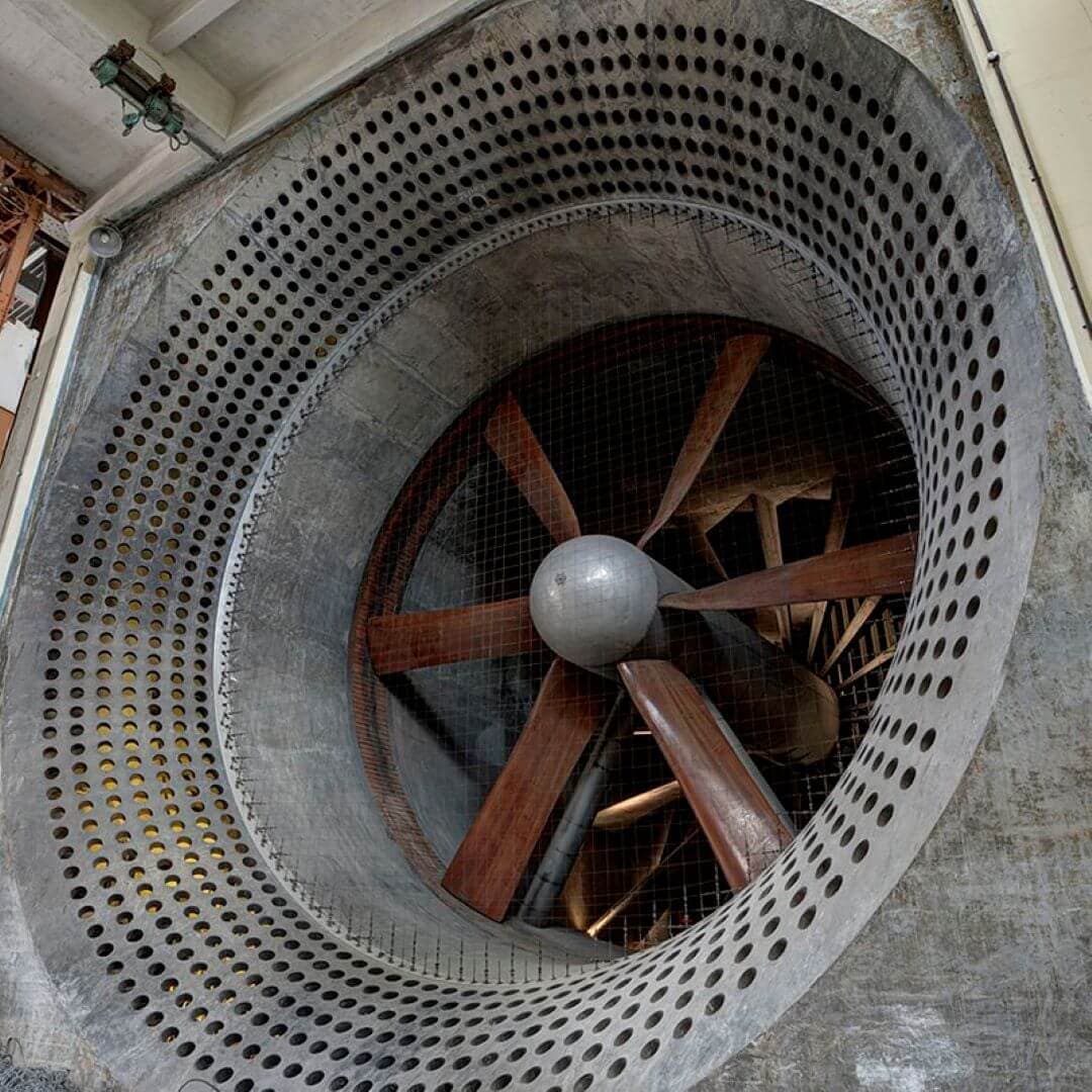 Large industrial fan with wooden blades inside a perforated metal casing, visible from a front-facing angle. - Home Instead