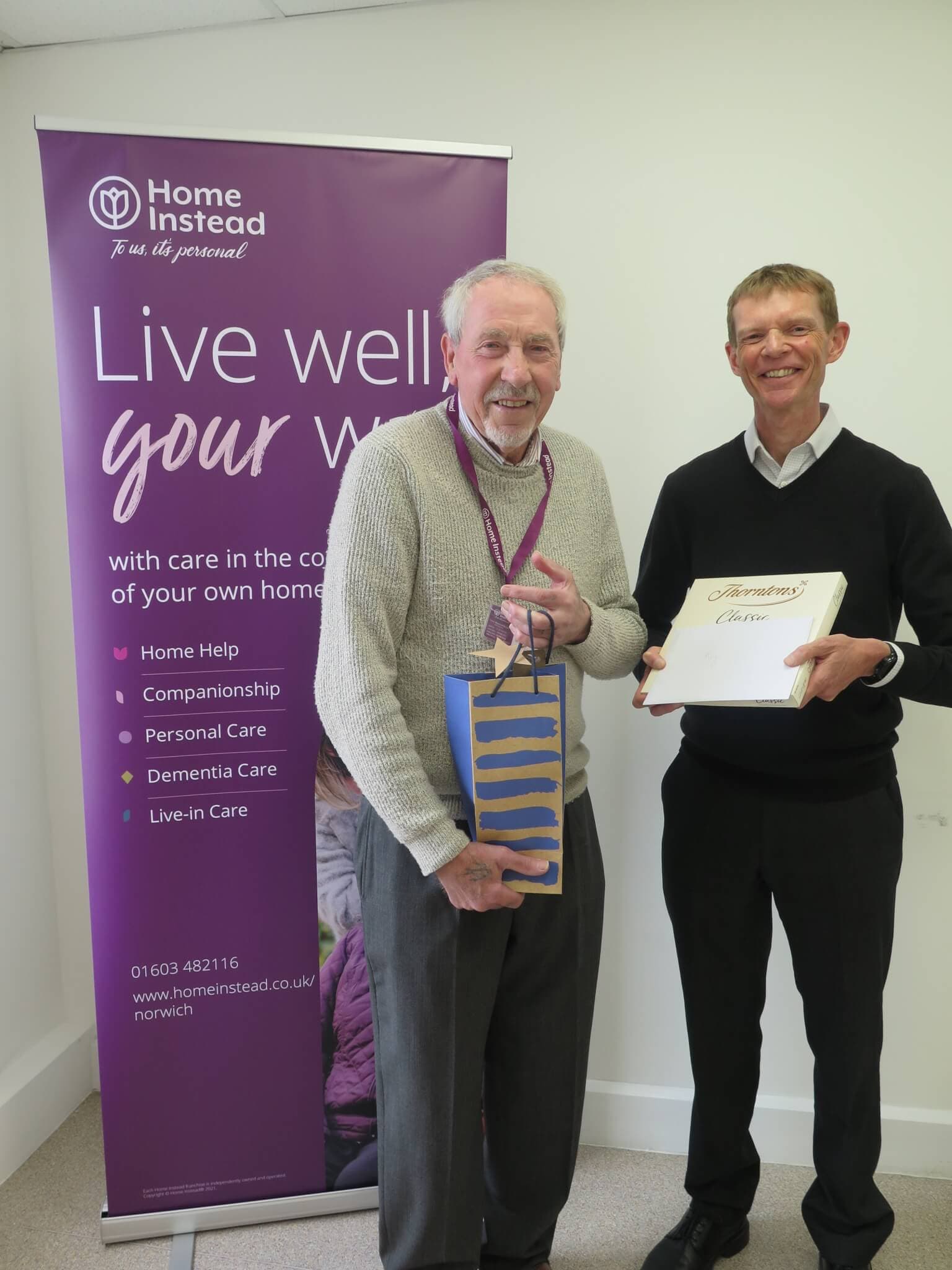 Two men stand smiling; one holds a certificate and the other holds a gift, next to a Home Instead banner. - Home Instead