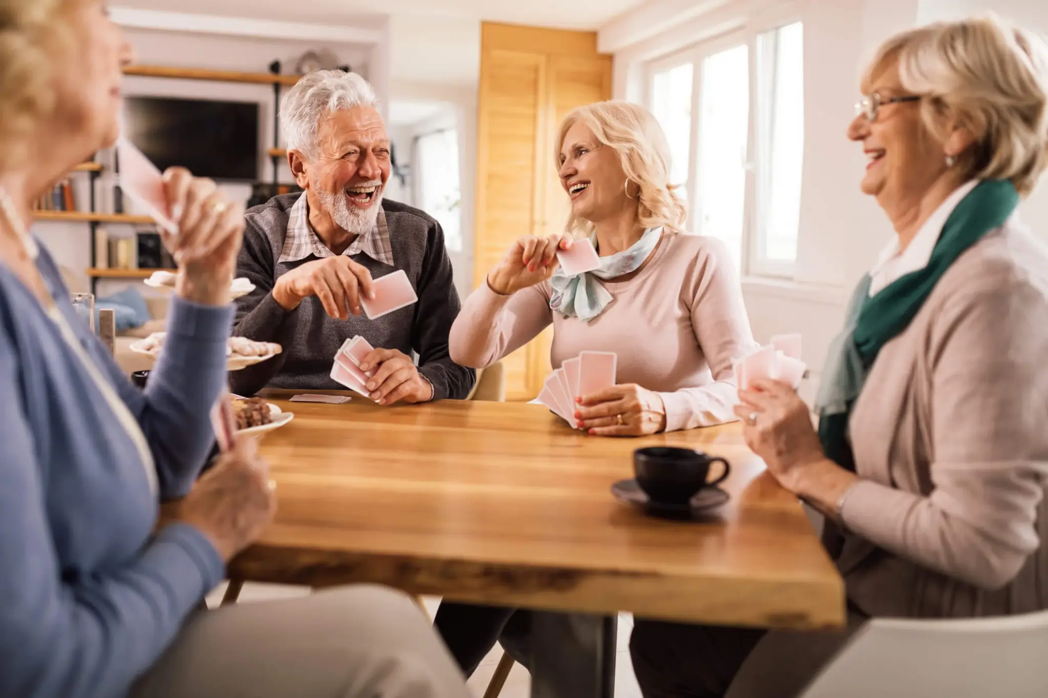 Four seniors joyfully playing cards at a wooden table in a cozy room, with coffee cups and snacks on the table. - Home Instead