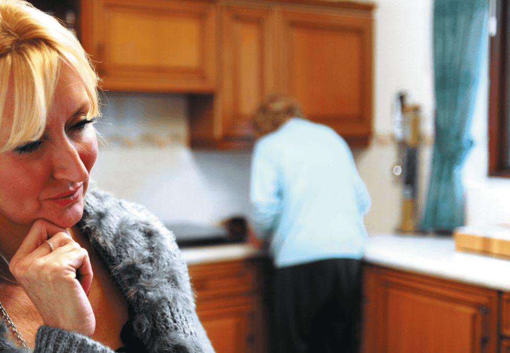 Woman in the foreground thinking, while another person with their back turned is in the kitchen. - Home Instead
