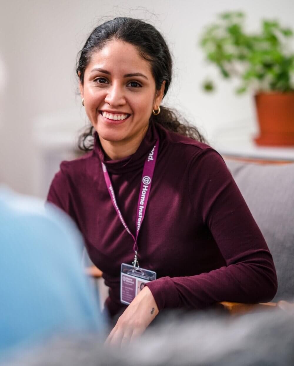 A woman with a name badge and lanyard smiles while sitting and talking in a cozy room with a plant in the background. - Home Instead