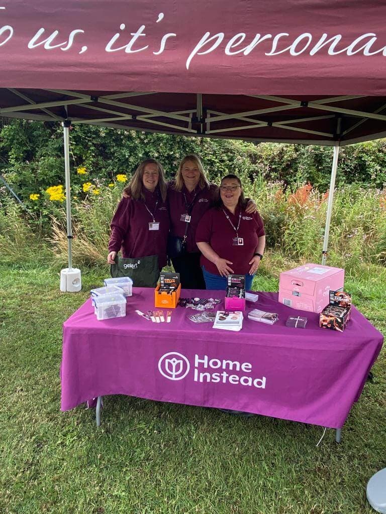Three people at a Home Instead booth with informational materials and promotional items under a tent outdoors. - Home Instead
