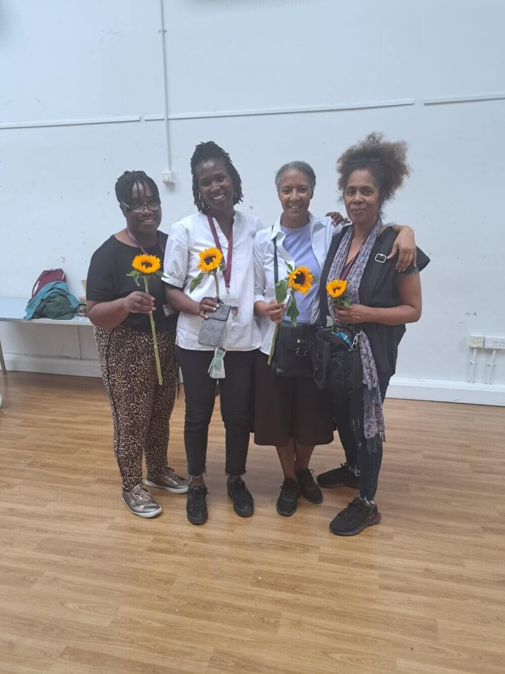 Four people smiling and holding sunflowers while standing on a wooden floor in a light-colored room. - Home Instead