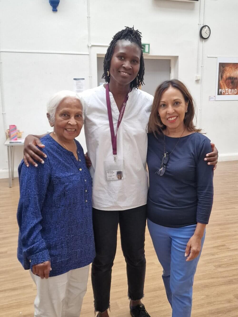 Three smiling women standing arm-in-arm in a room with wooden floors and white walls. - Home Instead
