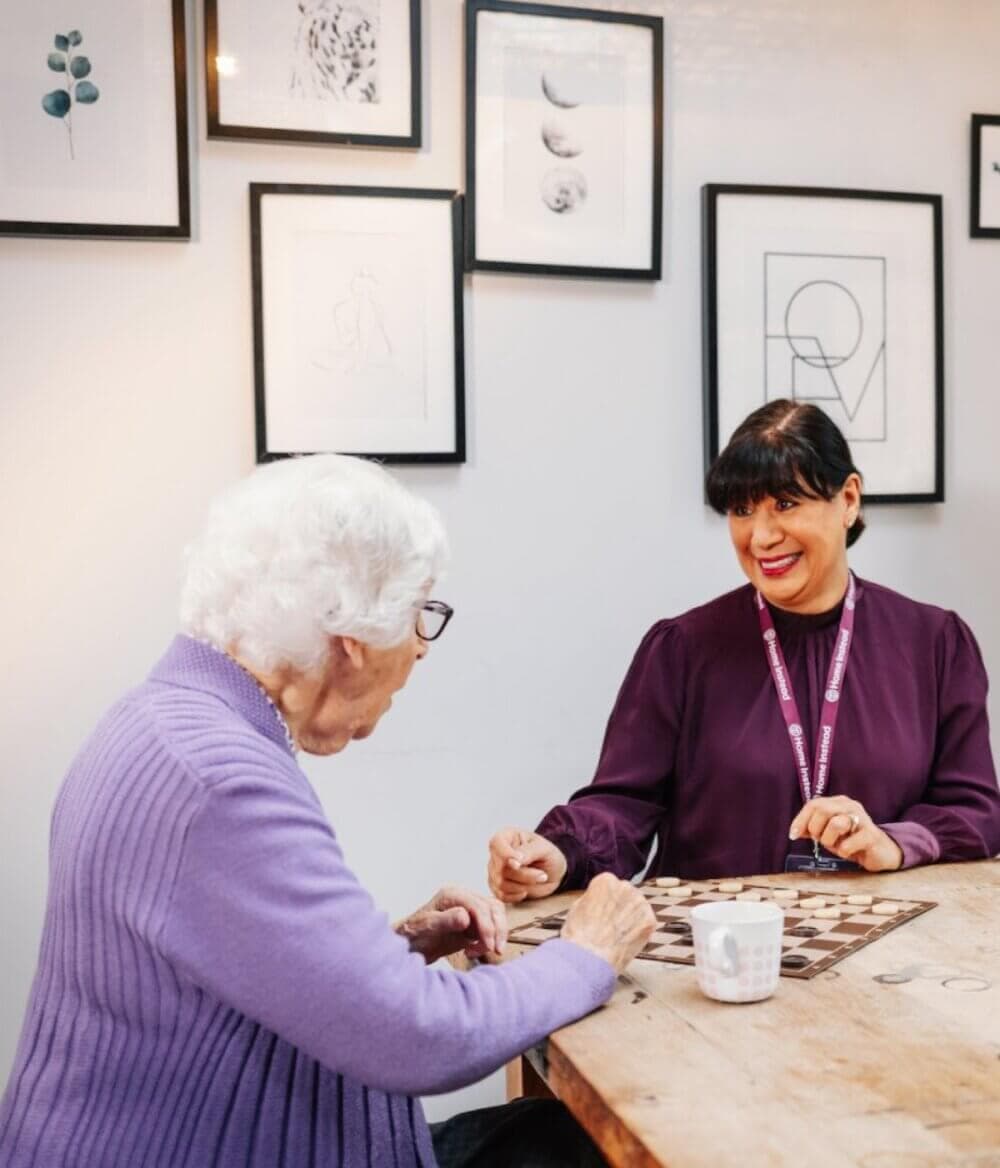 An elderly woman and a caregiver share a moment over a chessboard, surrounded by framed art on the wall. - Home Instead