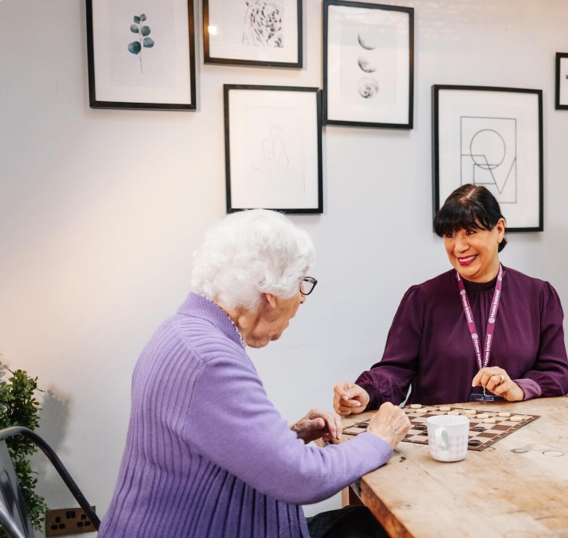An elderly woman and a younger woman sit at a table playing a board game, smiling in a room with framed art on the wall. - Home Instead