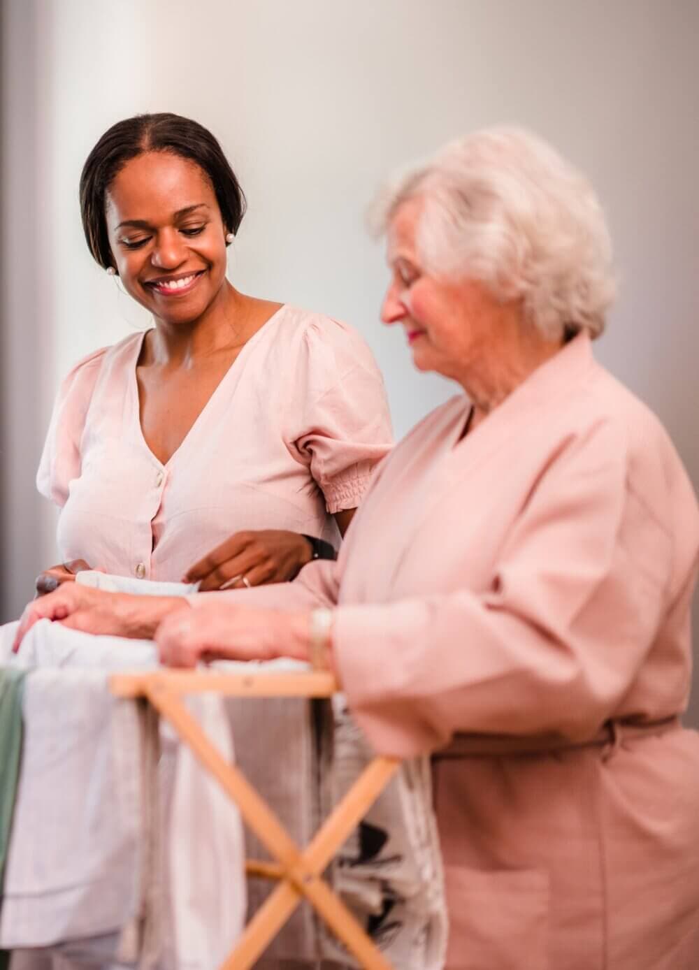 Two women in robes fold laundry together, one young and one elderly, both smiling warmly. - Home Instead