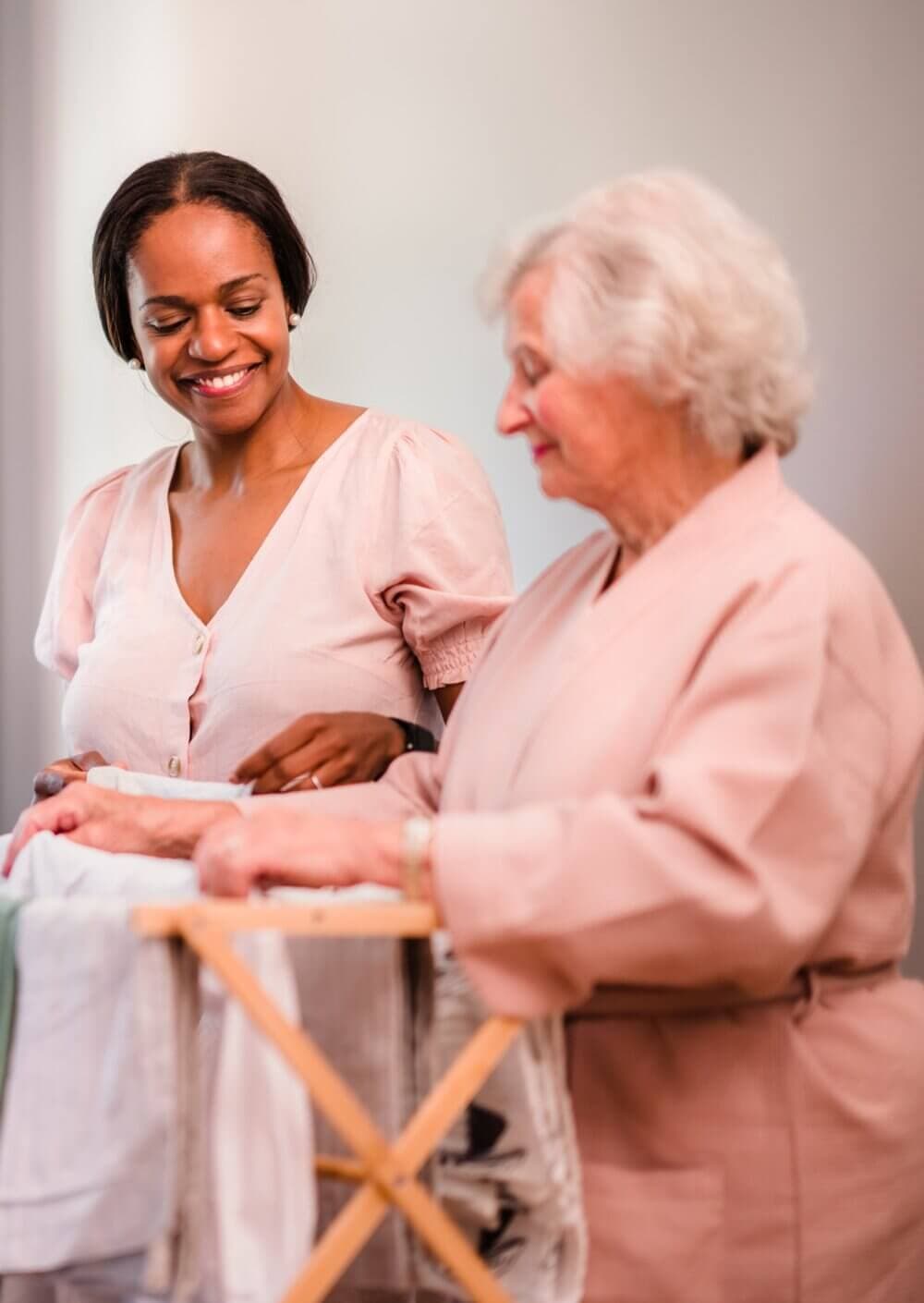A caregiver smiles while assisting an elderly woman folding laundry. - Home Instead