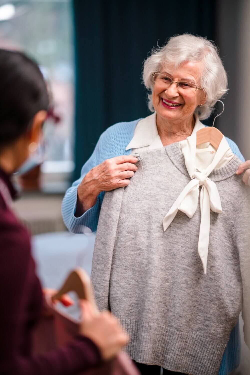 Elderly woman smiling while showing a gray sweater to another person holding a hanger. - Home Instead