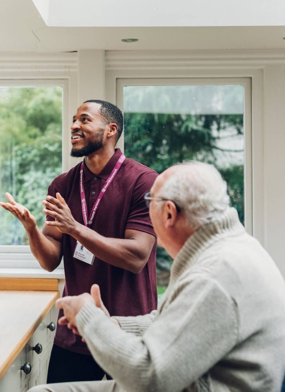 A young man in a maroon polo shirt engages in conversation with an elderly man in a gray sweater in a bright room. - Home Instead
