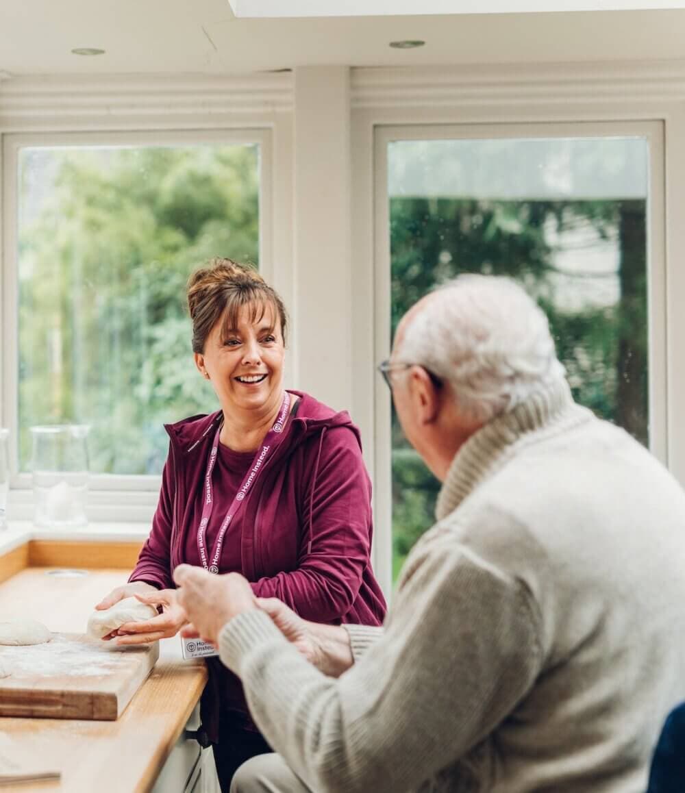 A woman applies makeup to an elderly person sitting in a chair inside a well-lit room with large windows. - Home Instead