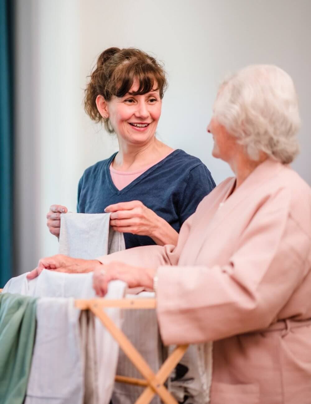 Two women conversing while folding laundry; one is older and seated, the other is younger and standing. - Home Instead Southampton
