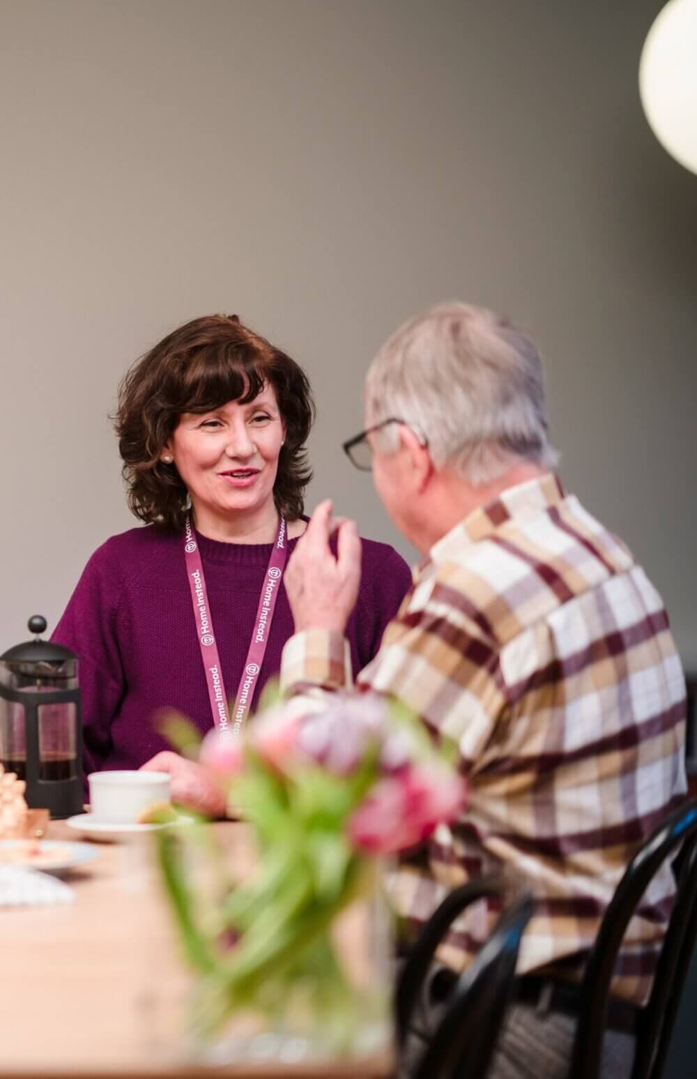 A woman and an older man having a conversation at a table, a bouquet of flowers in the foreground. - Home Instead