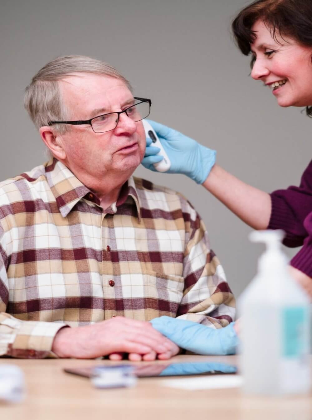 A woman uses a swab to clean an elderly man's cheek. He wears glasses and a plaid shirt, and she wears gloves and smiles. - Home Instead