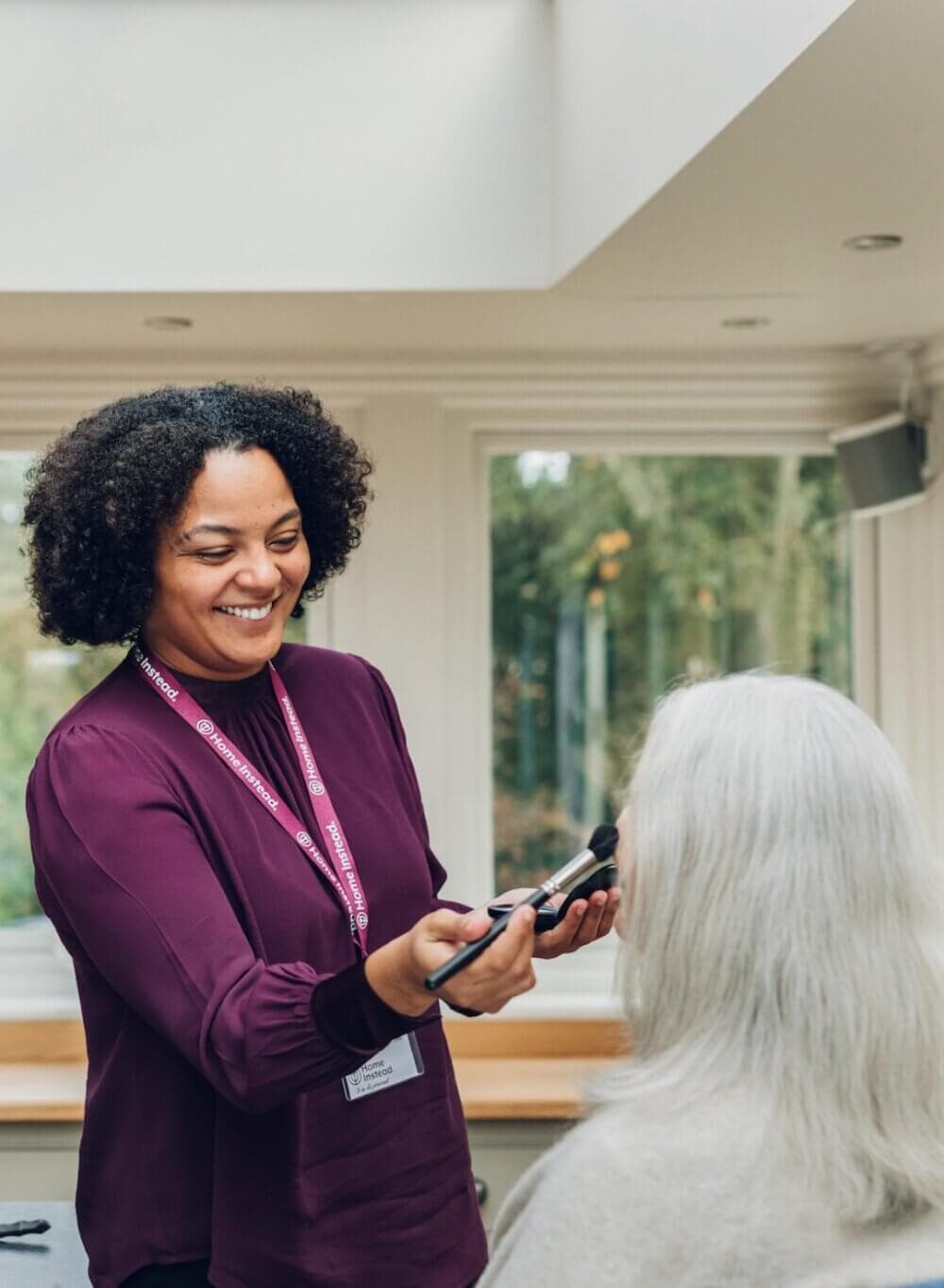 A smiling woman applies makeup to the face of an elderly person with white hair in a well-lit room. - Home Instead