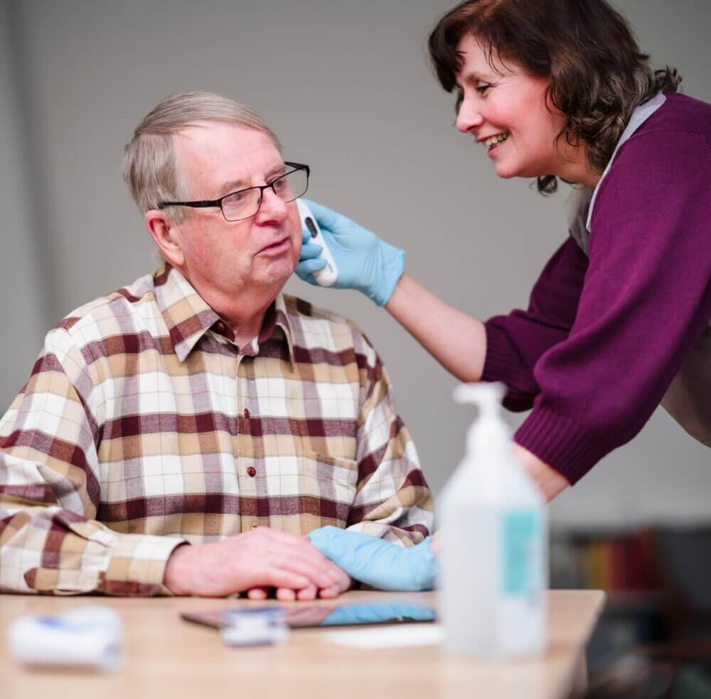 A woman in a purple blouse applies makeup to another person seated and facing away in a well-lit room. - Home Instead