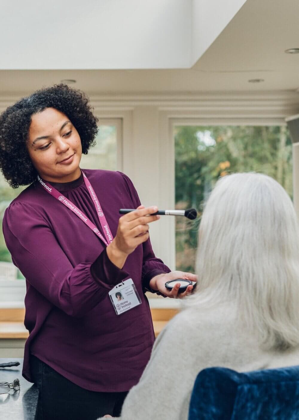 A woman with curly hair applies makeup to an elderly person with white hair at home. - Home Instead