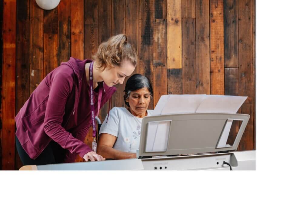 A woman with curly hair applies makeup to an older woman with long white hair, sitting in a bright room. - Home Instead