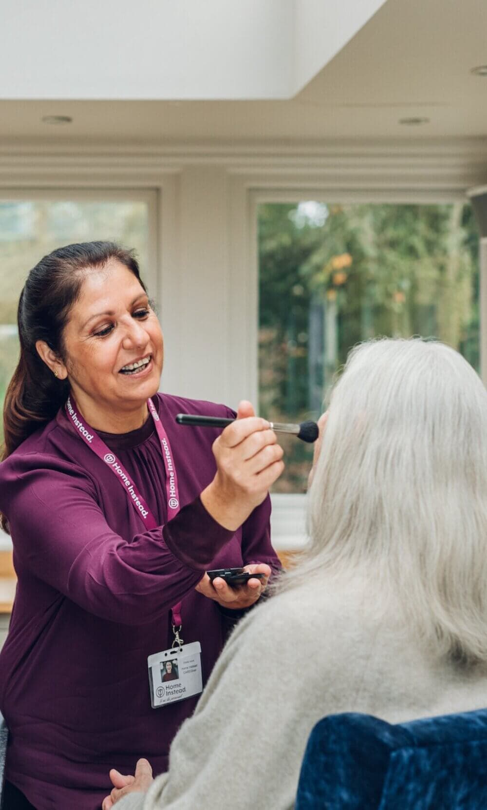 A woman smiles while applying makeup with a brush to an older woman sitting in a chair. - Home Instead