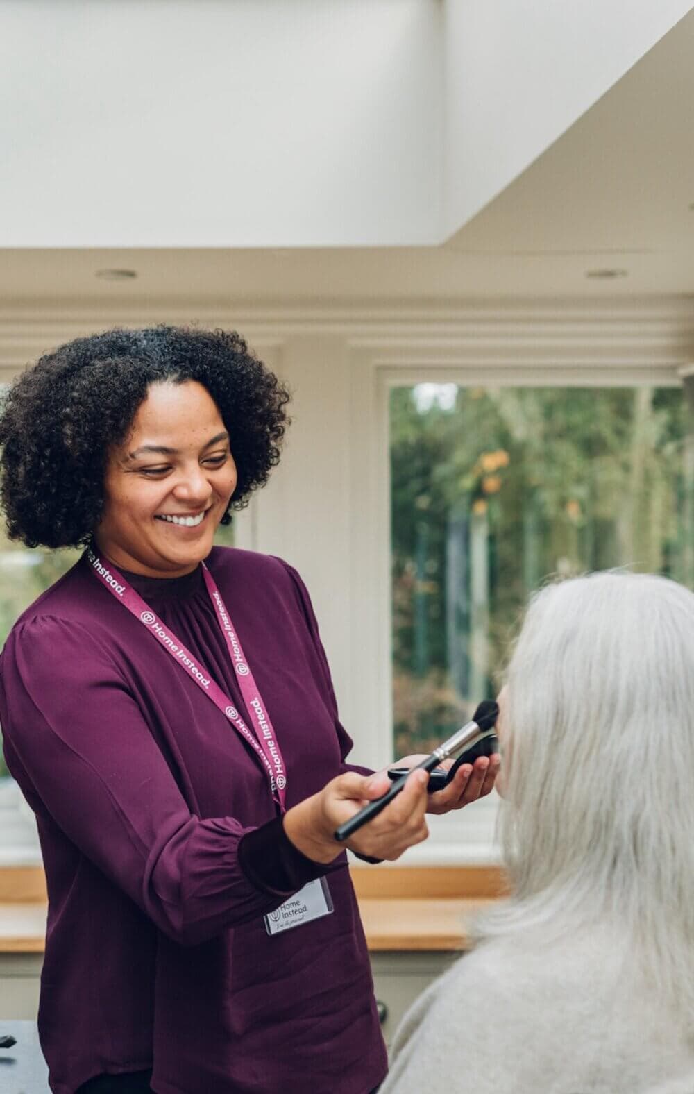 A woman wearing a badge joyfully applies makeup to an elderly woman in a well-lit room with large windows. - Home Instead