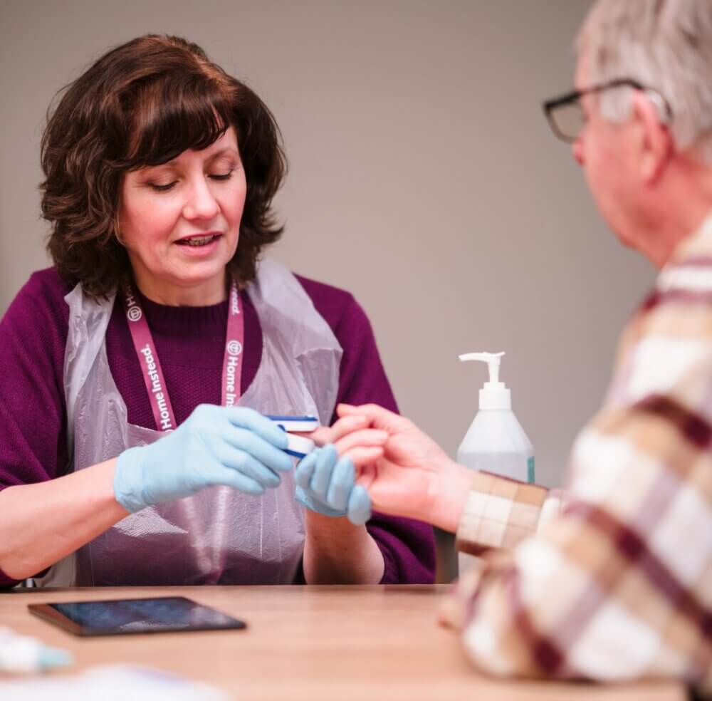 A healthcare worker in gloves and a plastic apron checks a man's finger, with a tablet and sanitizer on the table. - Home Instead