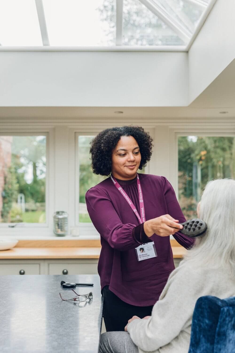 A woman wearing a purple top brushes the hair of a seated elderly person in a bright room with large windows. - Home Instead